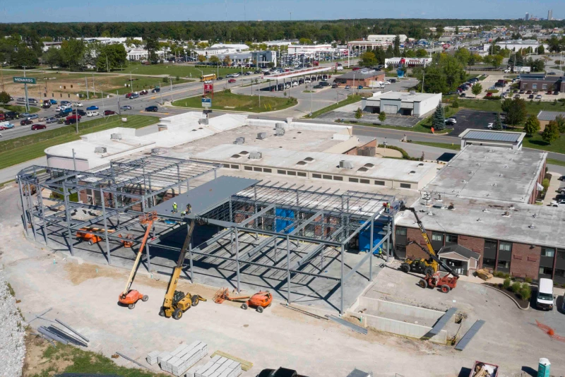 A high-angle shot of a new industrial or manufacturing facility under construction. The steel frame of the building is visible, with workers on a lift, installing structural components. The surrounding area includes other buildings, a road, and a parking lot.